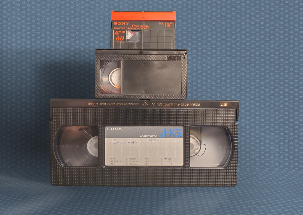 A photo of three video cassettes stacked on top of each other in front of a dark blue background.
The first tape is a MiniDV with a red tape cover flap. The second, in the middle, is a VHS-C.
The bottom tape is a standard VHS cassette.